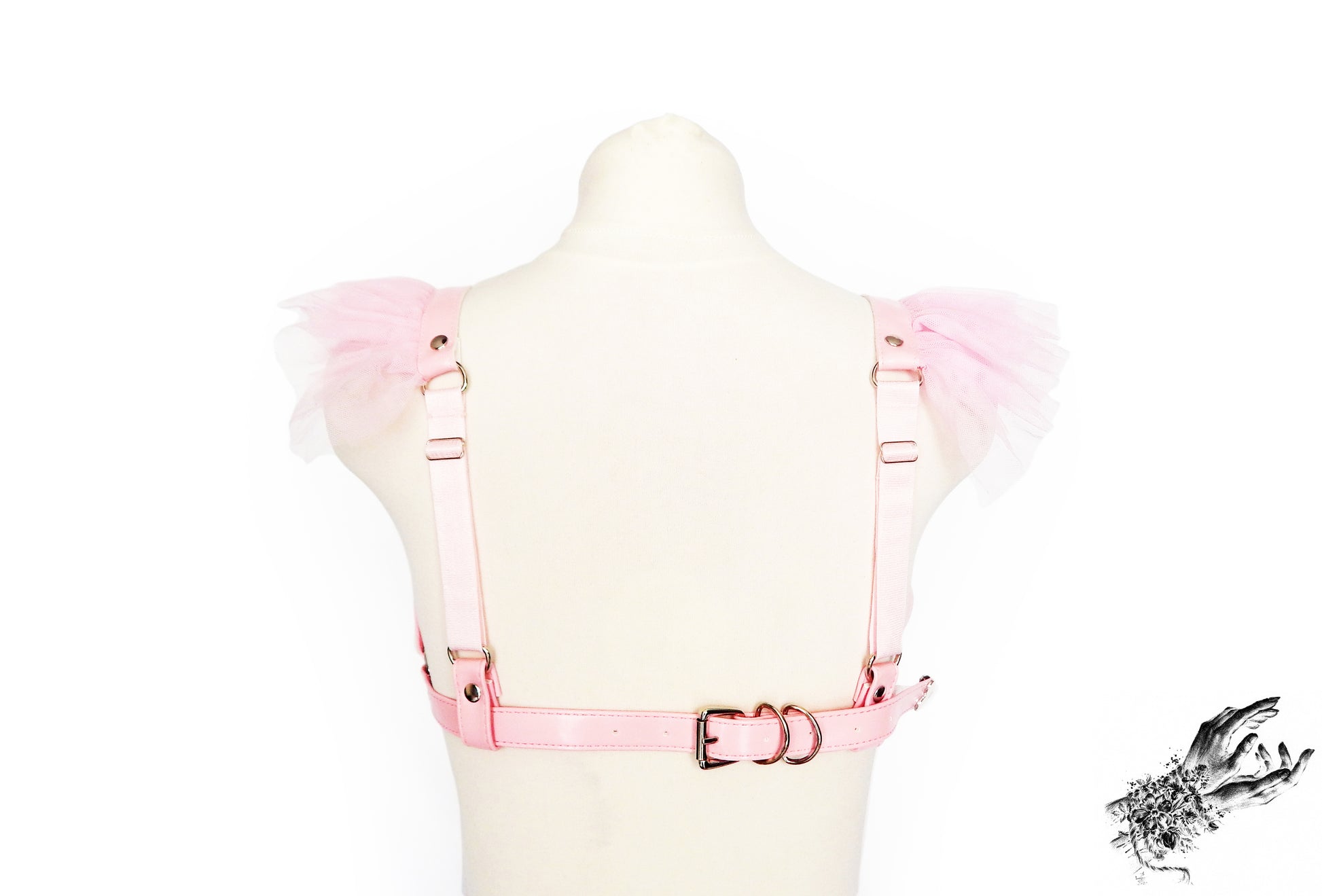 A light pink vegan leather harness bra with ruffled tulle accents on the shoulders and adjustable elastic shoulder straps and buckled faux leather chest strap, displayed on a mannequin against a white background.