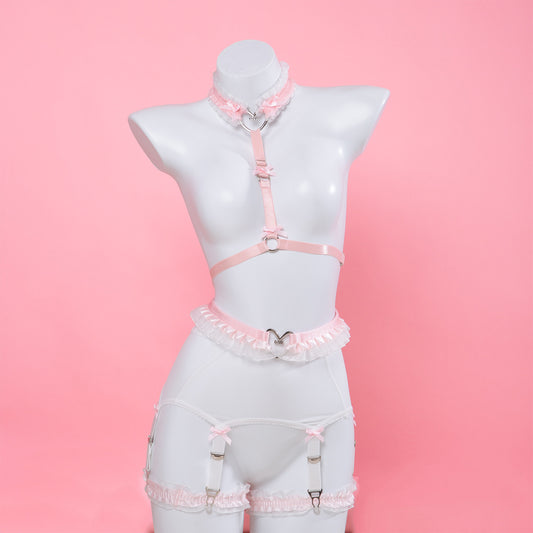 A pink and white lingerie set displayed on a white mannequin torso against a solid pink background. The set includes a pink and white collar with central silver heart ring, a pink halter-style harness with waist straps, a white and pink garter belt with lace trim, and matching pink and white ruffled thigh garters.