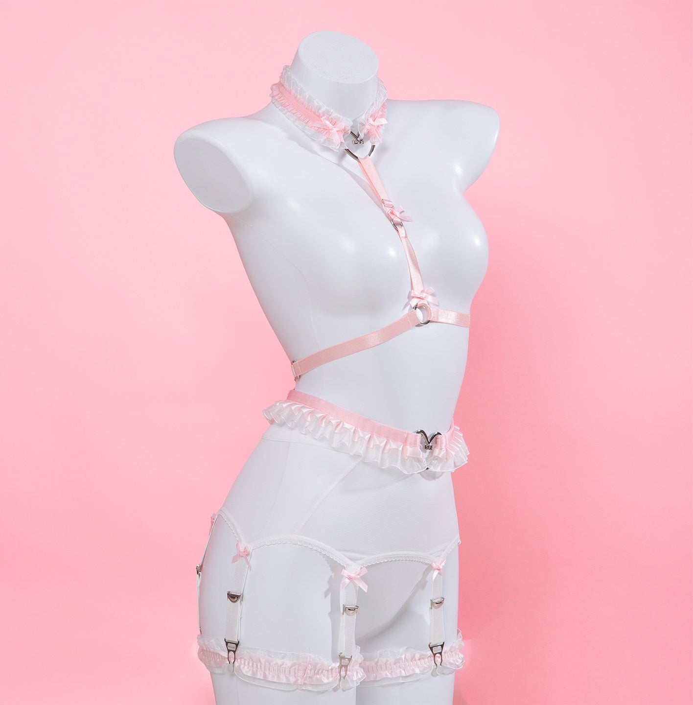 A pink and white strappy lingerie set displayed on a white female mannequin torso against a solid pink background. The set includes a choker collar, a harness-style top, a garter belt, and matching suspender belt with lace ruffles and silver-tone metal hardware.