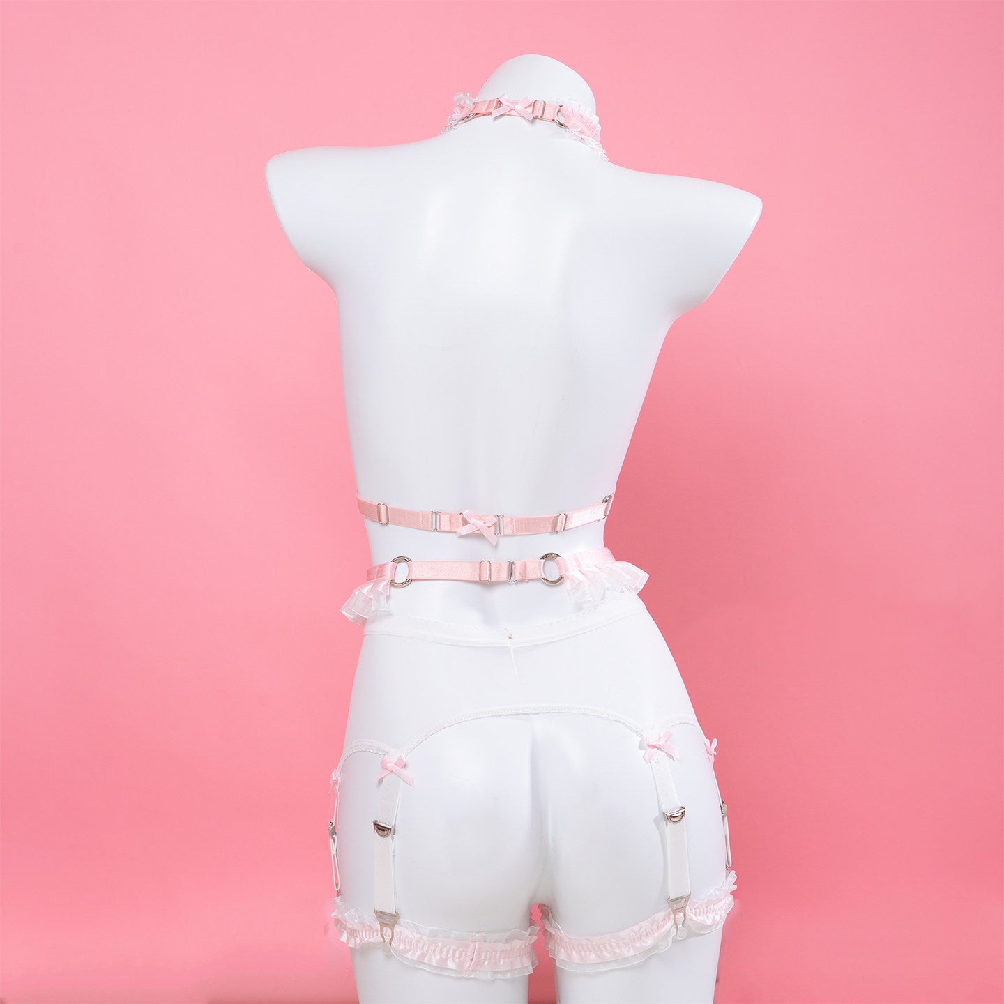 Pink lace and strap lingerie set displayed on a white mannequin from the back, featuring a ruffled choker, waist belt, and garter-style leg straps against a solid pink background.