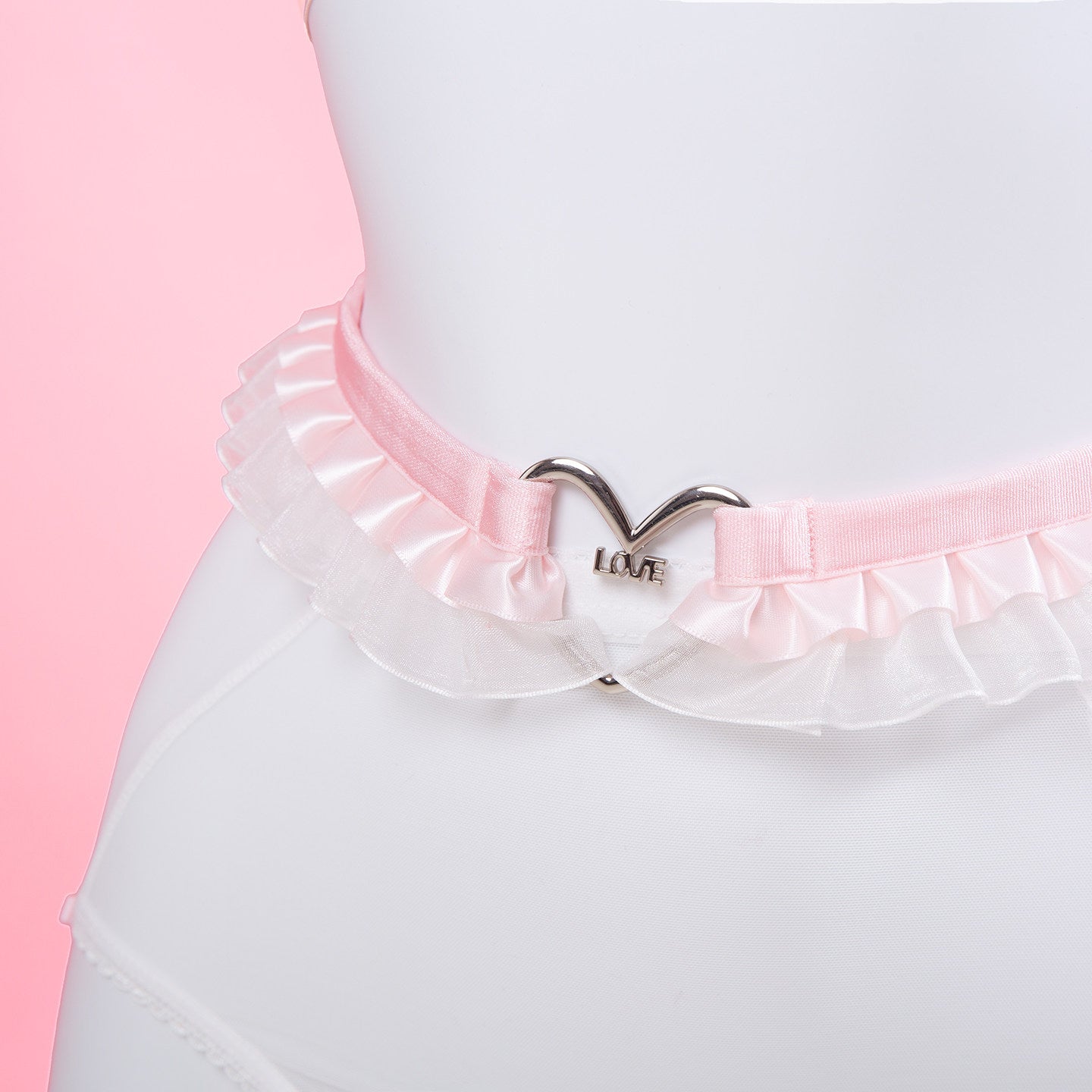 A light pink ruffled waist belt with a silver metal heart-shaped clasp engraved with the word 'LOVE', displayed on a white mannequin torso against a pink background.