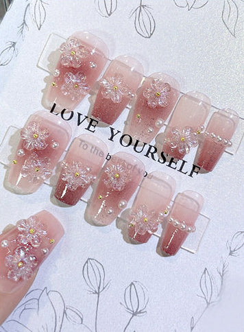 nude and pink glitter false nails featuring 3D sculpted gel flowers and white pearl beads, displayed on a white backing card.