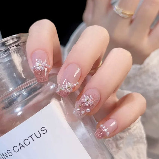 nude and pink glitter false nails featuring 3D sculpted gel flowers and white pearl beads, modelled on a hand holding a perfume bottle.