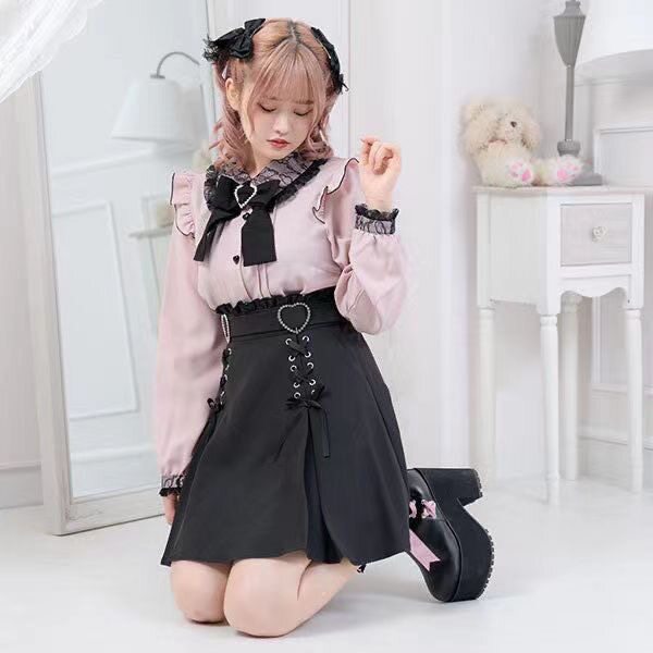 Modelled photo of a pink and black long sleeved blouse featuring a Peter Pan collar, ruffled shoulders, pintucks, heart shaped buttons and detachable bow brooch. Styled with a black ruffled skirt and boots for a Jirai Kei aesthetic.