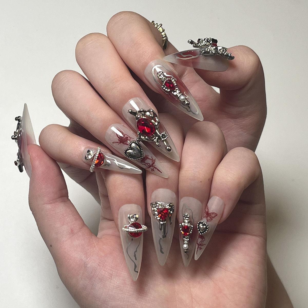 Long stiletto length false nails featuring white and red blended bases with silver swirls and flames, red butterflies, 3D silver and red crystal heart, butterfly and star charms, modelled on a pair of hands against a white background.