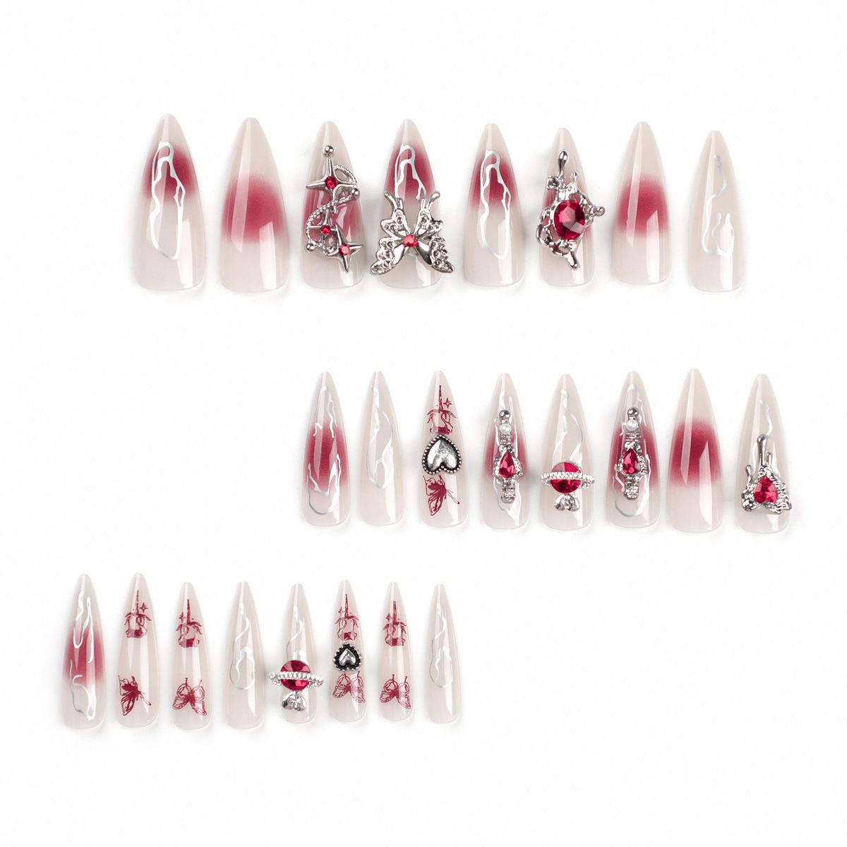 Three rows of pointed false press-on nails on a plain white background. The nails are glossy with a red-to-clear ombre gradient and various decorative embellishments including red gems, butterflies, hearts, stars and swirls.