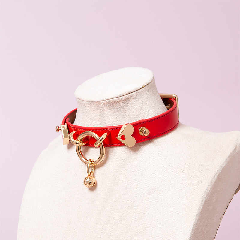 Red leather choker featuring a light gold tone central O ring, heart shaped hardare and detachable bell charm. Displayed on a white mannequin bust against a pink background for a kawaii aesthetic.
