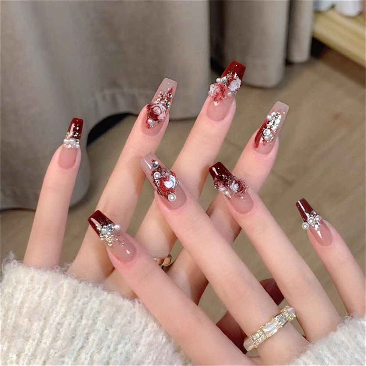 long ballerina shape press-on nails featuring a nude and red base, 3D white and red bloody rose charms, pearls and diamantes, modelled on hands wearing a ring and fluffy sweater.