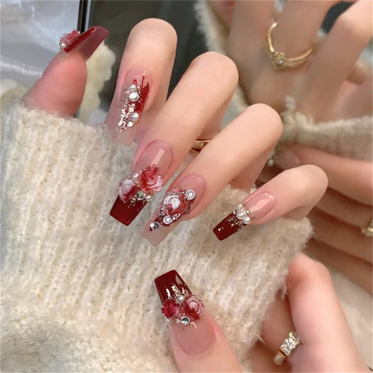 long square press-on nails featuring a nude and red base, 3D white and red bloody rose charms, pearls and diamantes, modelled on a hand resting on a fluffy sweater.