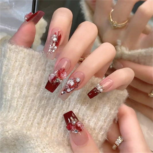 long square press-on nails featuring a nude and red base, 3D white and red bloody rose charms, pearls and diamantes, modelled on a hand resting on a fluffy sweater.