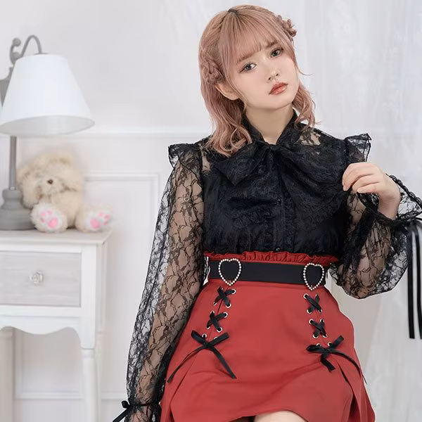 Model photo of a red high waisted skirt featuring a ruffled waist with side zip fastening, diamante heart shaped buckle belt, black satin ribbon corset lacing and pleated a-line design, styled with a black ruffled blouse for a Jirai Kei fashion aesthetic.