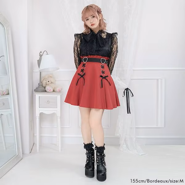 Modelled photo of a red high waisted skirt featuring a ruffled waist, diamante heart shaped buckle belt, black satin ribbon corset lacing and pleated a-line design, styled with a black blouse and chunky black Mary Jane shoes for a Jirai Kei fashion aesthetic.