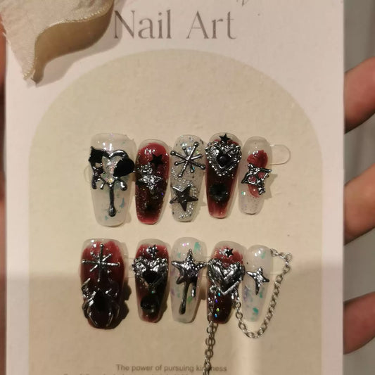 A set of decorative press-on nails displayed on a card, with a hand holding the card. The nails feature dark red and clear designs with silver glitter, metallic starbursts, and silver chain details.