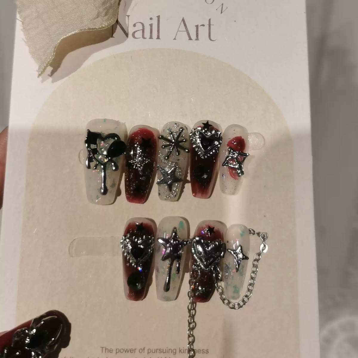 A set of decorative press-on nails displayed on a card, with a hand holding the card. The nails feature dark red and clear designs with silver glitter, metallic starbursts, and silver chain details.