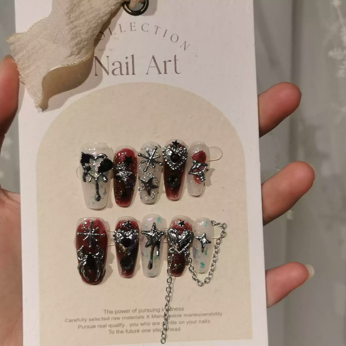 A set of decorative press-on nails displayed on a card, with a hand holding the card. The nails feature dark red and clear designs with silver glitter, metallic starbursts, and silver chain details.