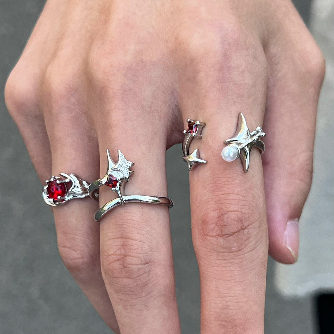 A silver tone ring featuring a starburst design, dark red crystal and white pearl, worn on a hand with two other matching rings.
