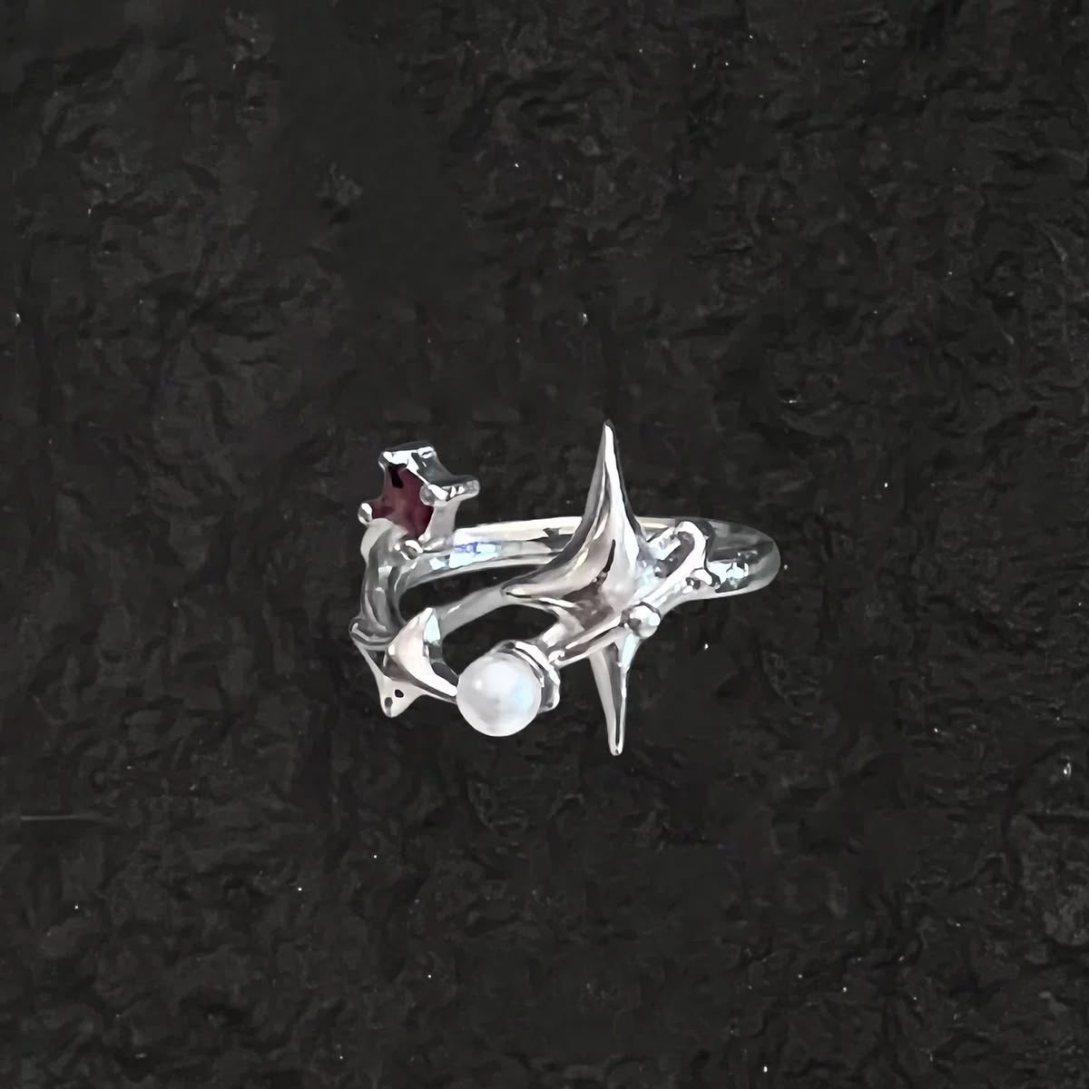A silver-toned gothic ring featuring a wraparound starburst design with a white pearl and a small dark red gemstone, set against a dark textured background.