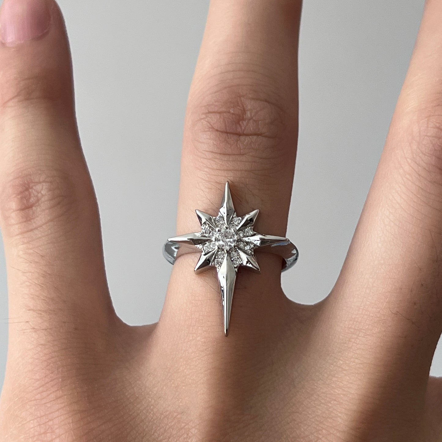 A silver-toned starburst ring with a central clear gemstone, shown worn on a finger against a soft light gray background.
