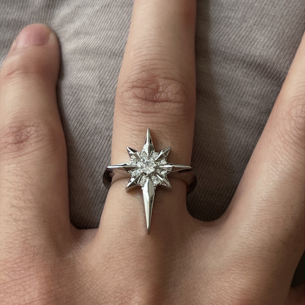 A silver-toned north star ring with clear central zircon gemstones, shown worn on a person's finger against a gray fabric background.