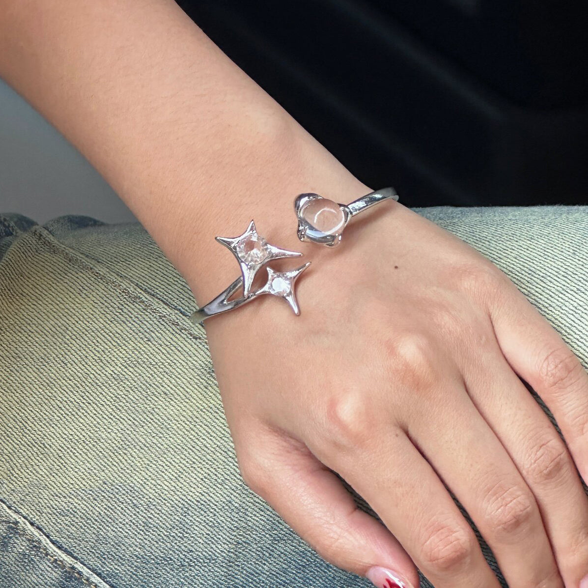Worn photo of a silver plated costume jewellery bangle featuring a silver starburst design, wrap-around cuff and clear round glass crystal gemstones, styled with faded denim jeans for a street fashion aesthetic.