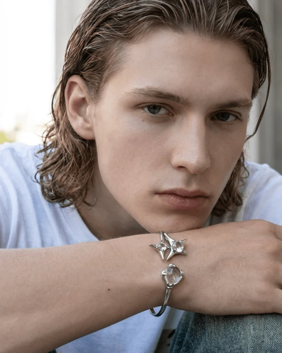 Modelled photo of a silver plated bangle featuring a silver starburst design, wrap-around cuff and clear round glass crystal gemstones, styled with slicked back, wet-look hair and a white t-shirt for an edgy street fashion style.
