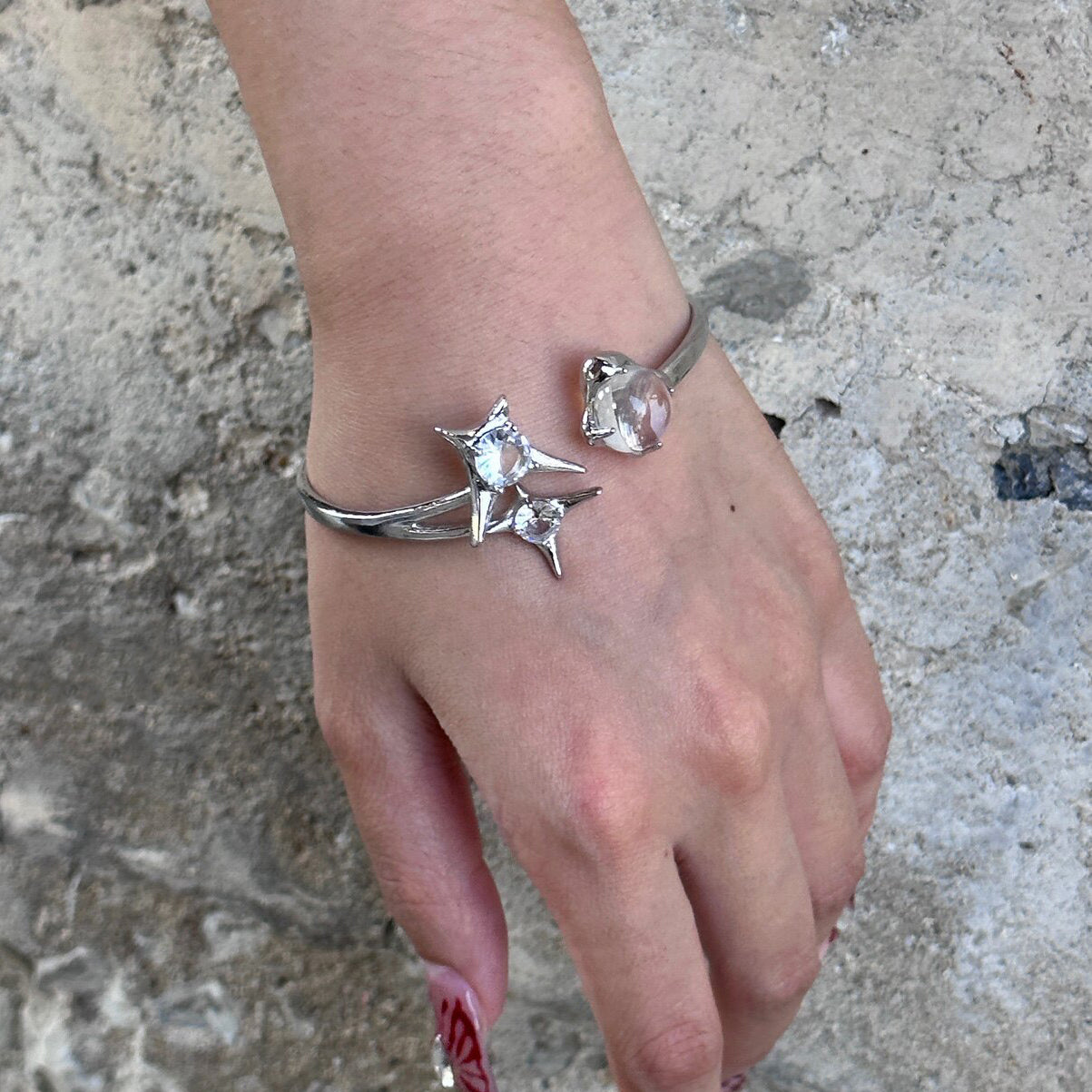 Worn photo of a silver plated costume jewellery bangle featuring a silver starburst design, wrap-around cuff and clear round glass crystal gemstones, styled with long red nails for a street fashion aesthetic.