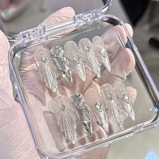 A hand wearing a light pink glove holds an open clear plastic case containing press-on nails. The nails are long stiletto-shaped with a sheer nude-pink base, heavy silver glitter, and large rhinestone embellishments.