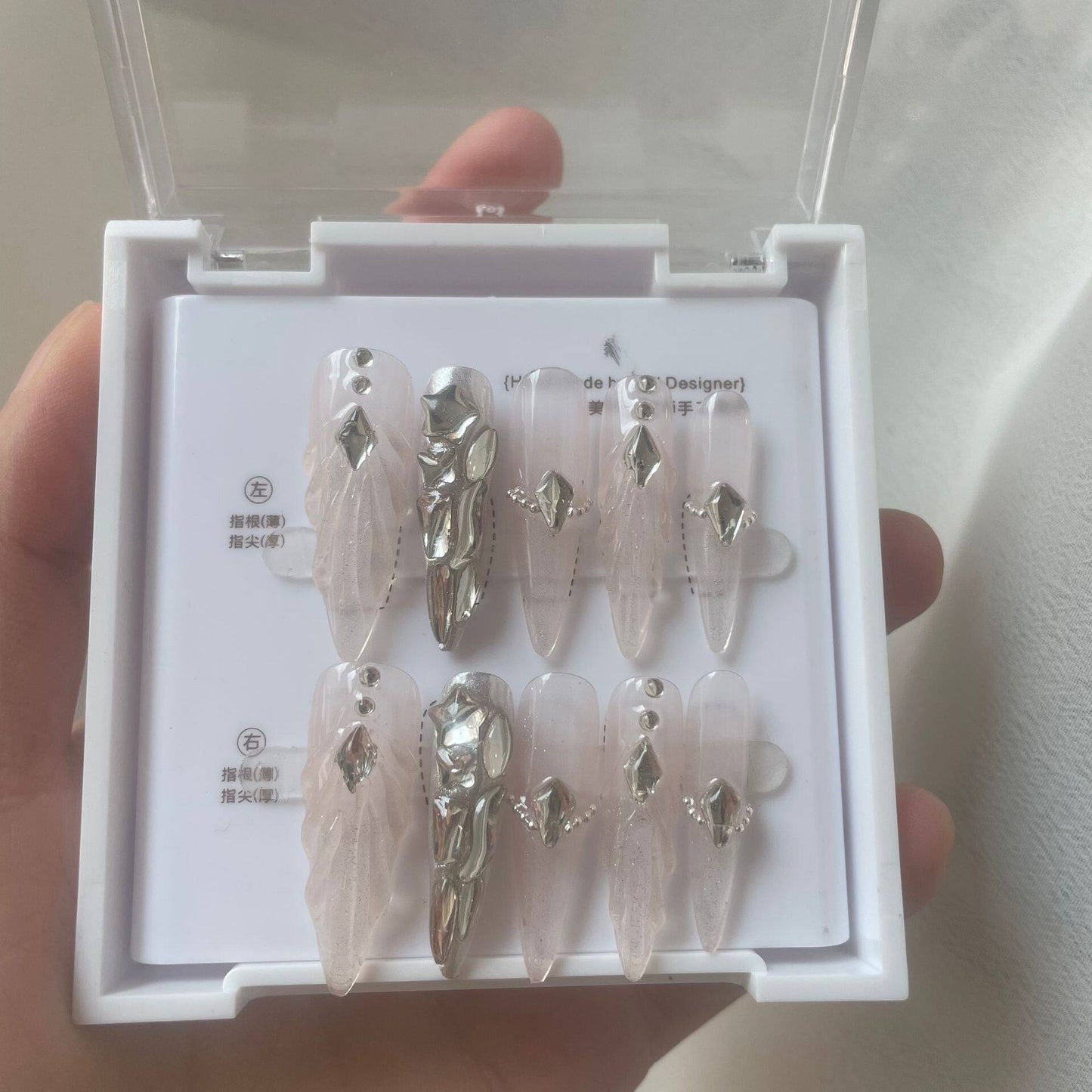 A hand holding a white plastic case containing a set of press-on nails. The nails are long and ballerina-shaped, featuring a sheer pale pink base with delicate sculpted scallops and ripples with shiny silver rhinestone embellishments and silver chrome accent nail.
