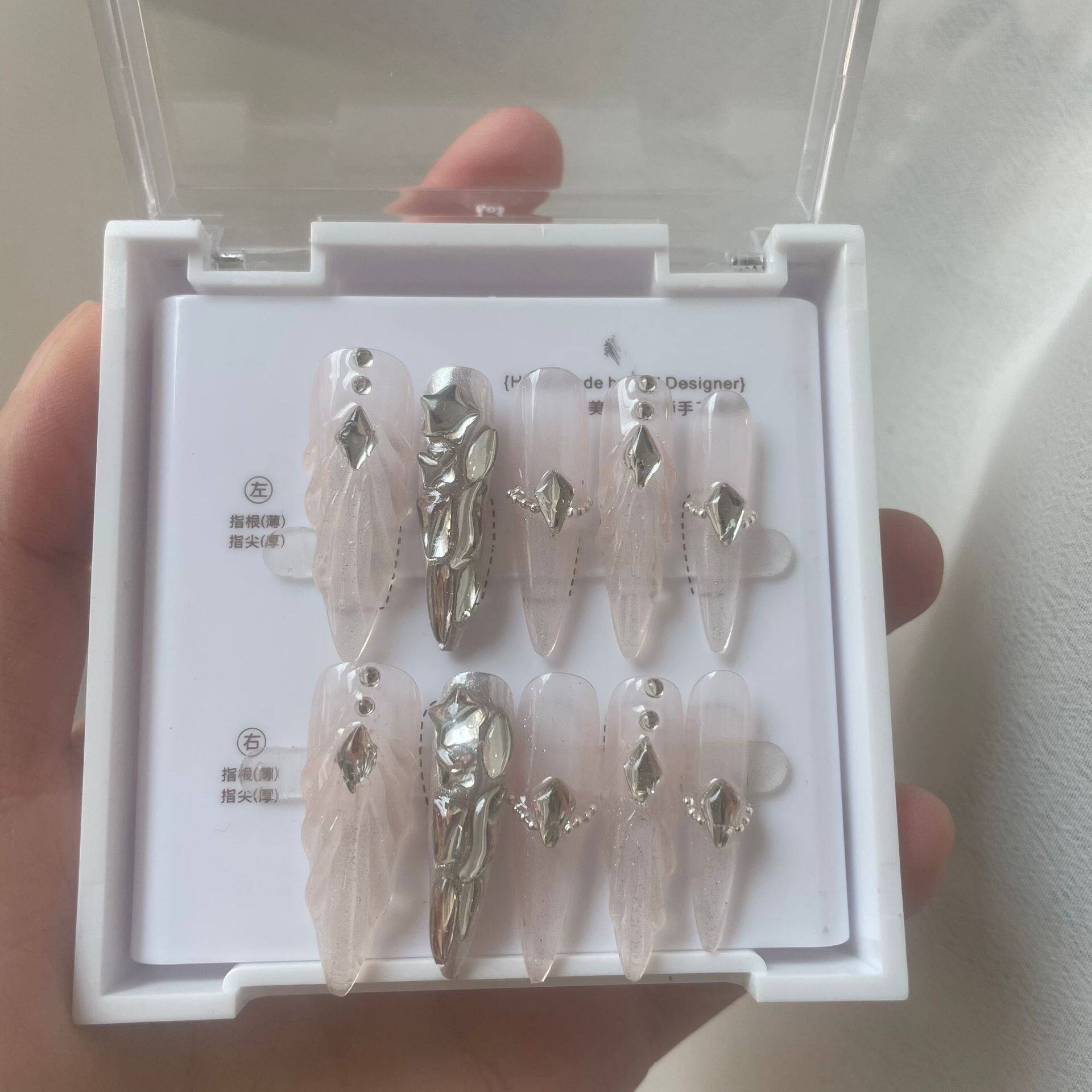A hand holding a white plastic case containing a set of press-on nails. The nails are long and ballerina-shaped, featuring a sheer pale pink base with delicate sculpted scallops and ripples with shiny silver rhinestone embellishments and silver chrome accent nail.
