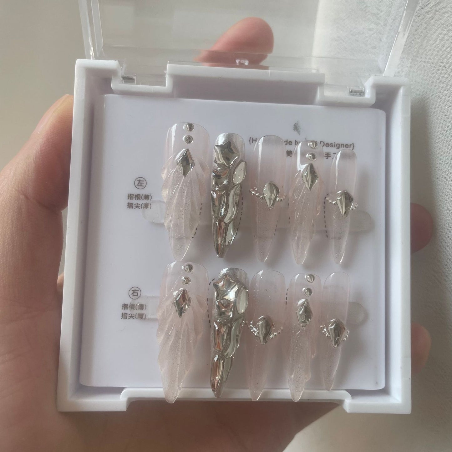 A hand holding a white plastic case containing a set of press-on nails. The nails are long and ballerina-shaped, featuring a sheer pale pink base with delicate sculpted scallops and ripples with shiny silver rhinestone embellishments and silver chrome accent nail.