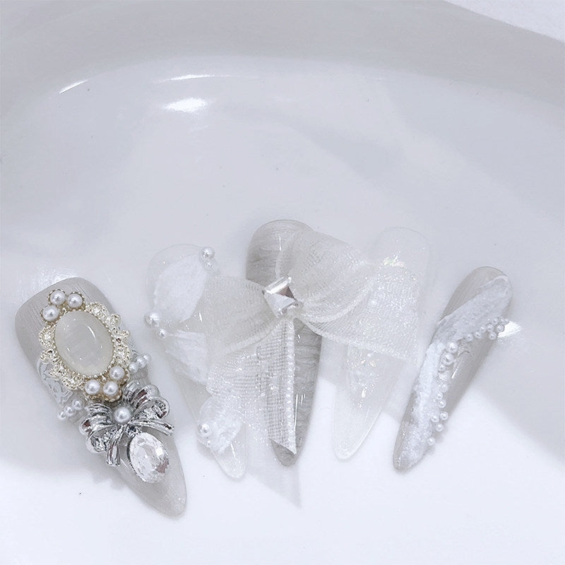 A set of decorative false nails in a bridal or Gyaru style, featuring sheer white organza bows, white faux pearls, silver rhinestone accents, and a large oval pearl, displayed on a white background.