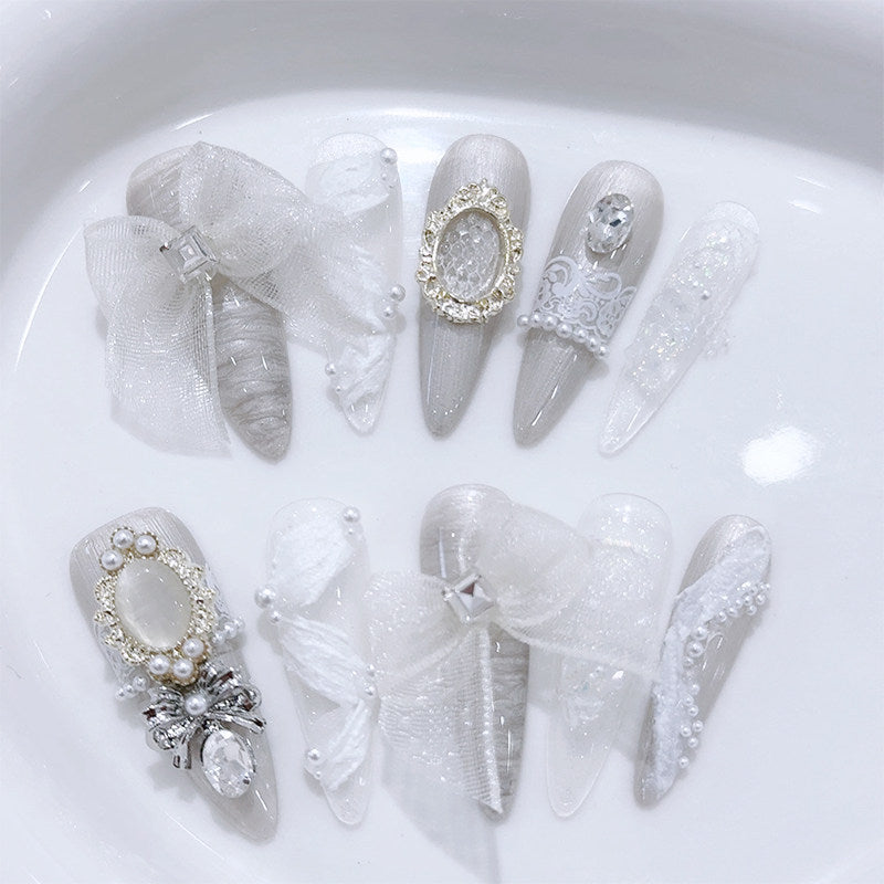 A set of white and silver decorative press-on nails arranged on a glossy white surface. The nails feature lace, sheer mesh, pearls, rhinestones, bows, and ornate embellishments in white and silver tones.