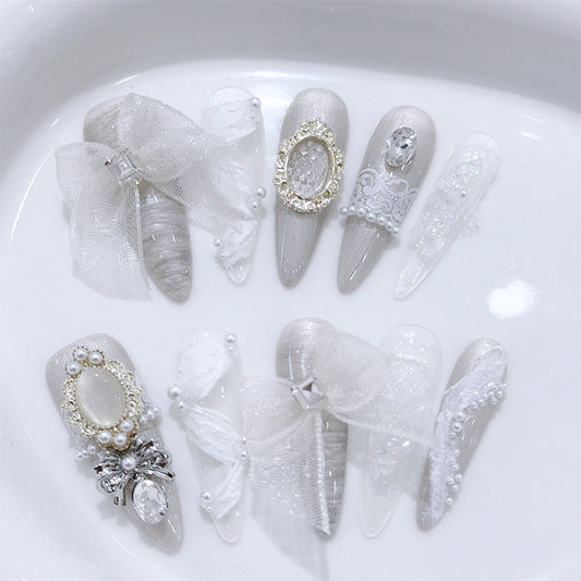 A set of white and silver decorative press-on nails arranged on a glossy white surface. The nails feature lace, sheer mesh, pearls, rhinestones, bows, and ornate embellishments in white and silver tones.