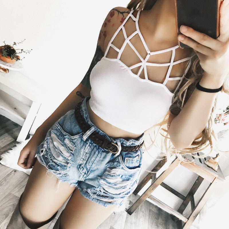 A woman wearing a white strappy crop top with an intricate geometric cage design on the back and light-wash distressed denim shorts, taking a mirror selfie.