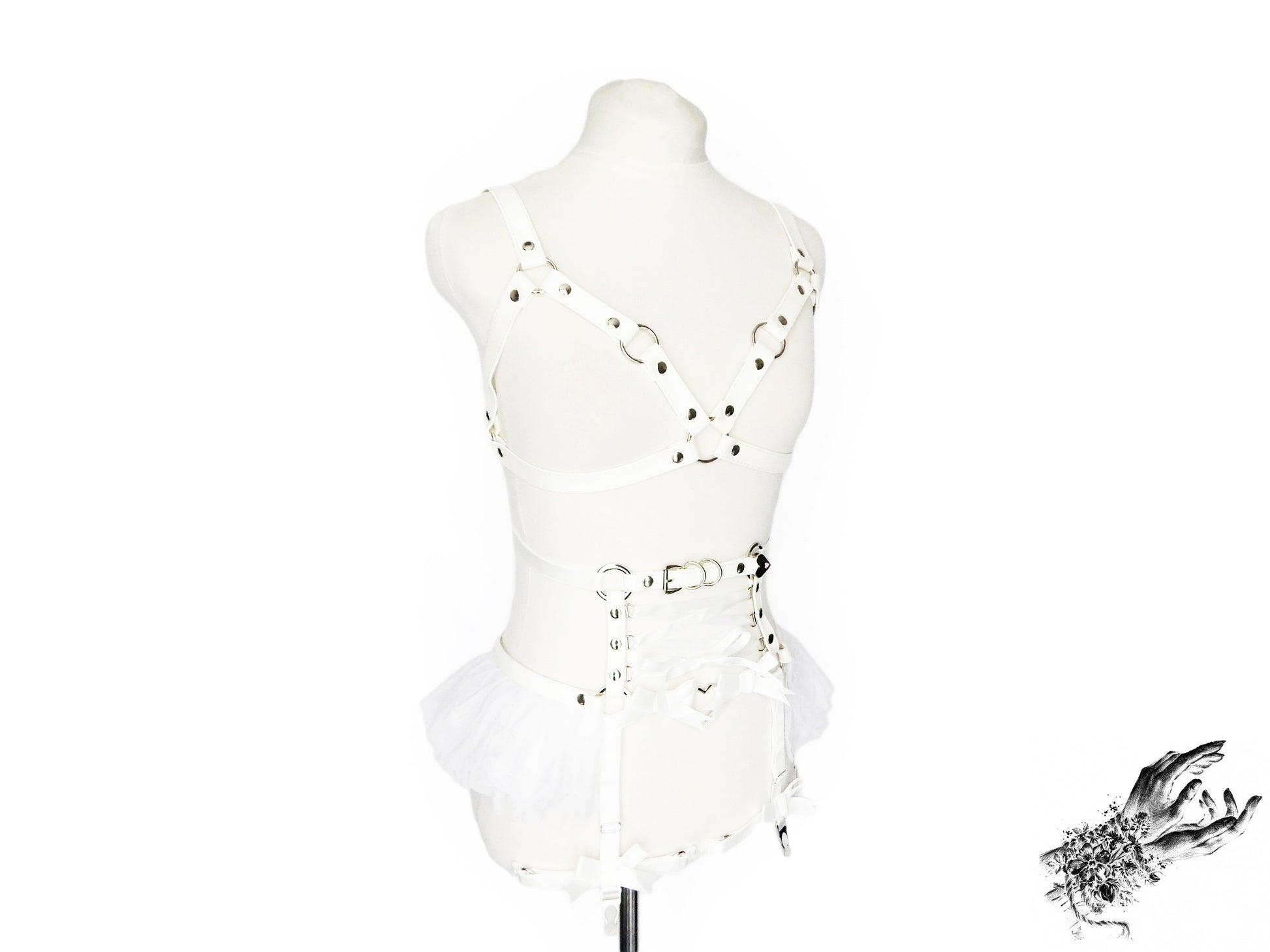 A white vegan leather harness set featuring a separate harness bra and garter belt with silver heart ring hardware, white ruffled tulle accents and corset lacing at the waist, displayed on a mannequin against a white background.