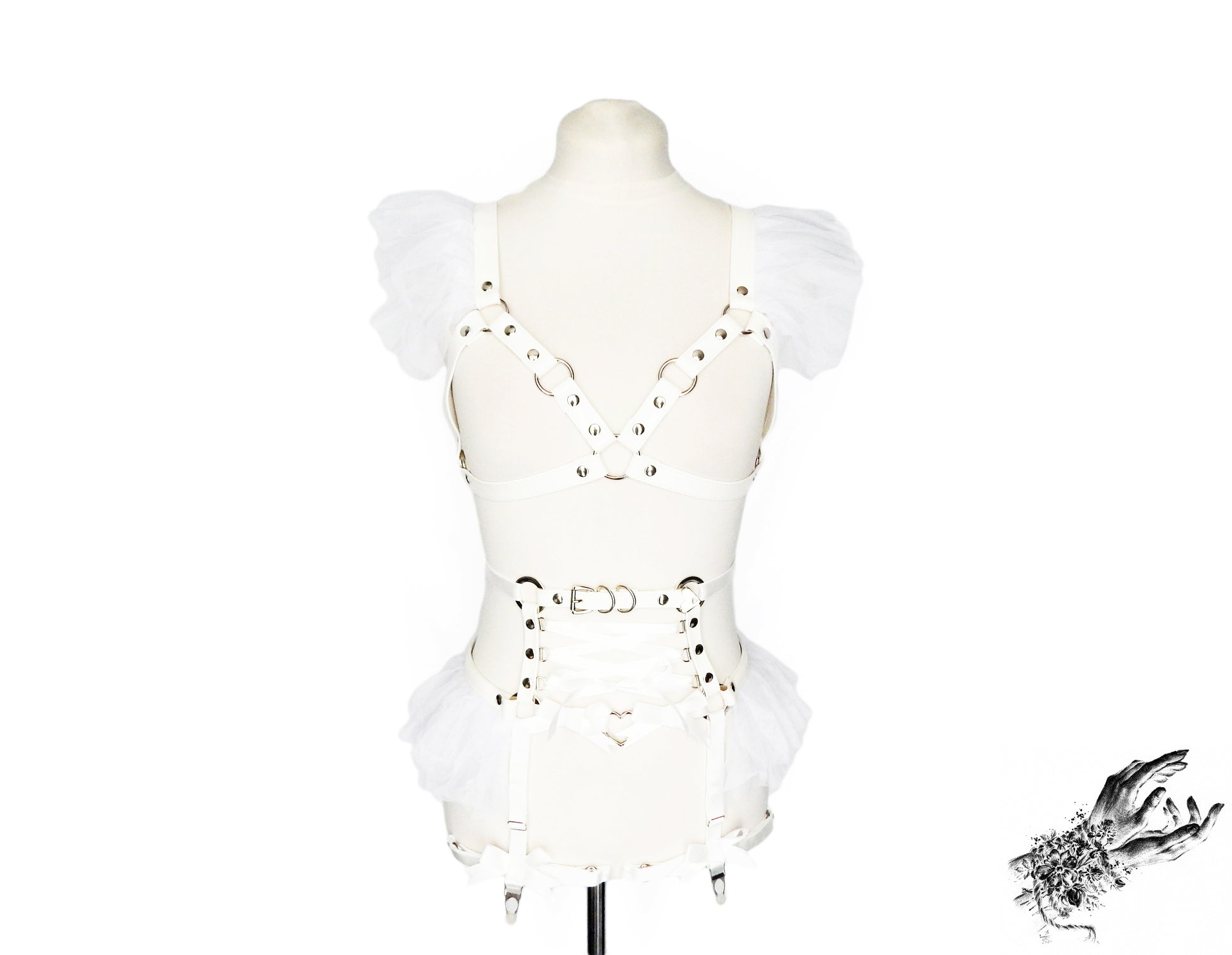 Front view of a white vegan leather harness set featuring a separate harness bra and garter belt with white ruffled tulle accents at the shoulders and hips, corset lacing and silver heart ring hardware, displayed on a mannequin against a white background.
