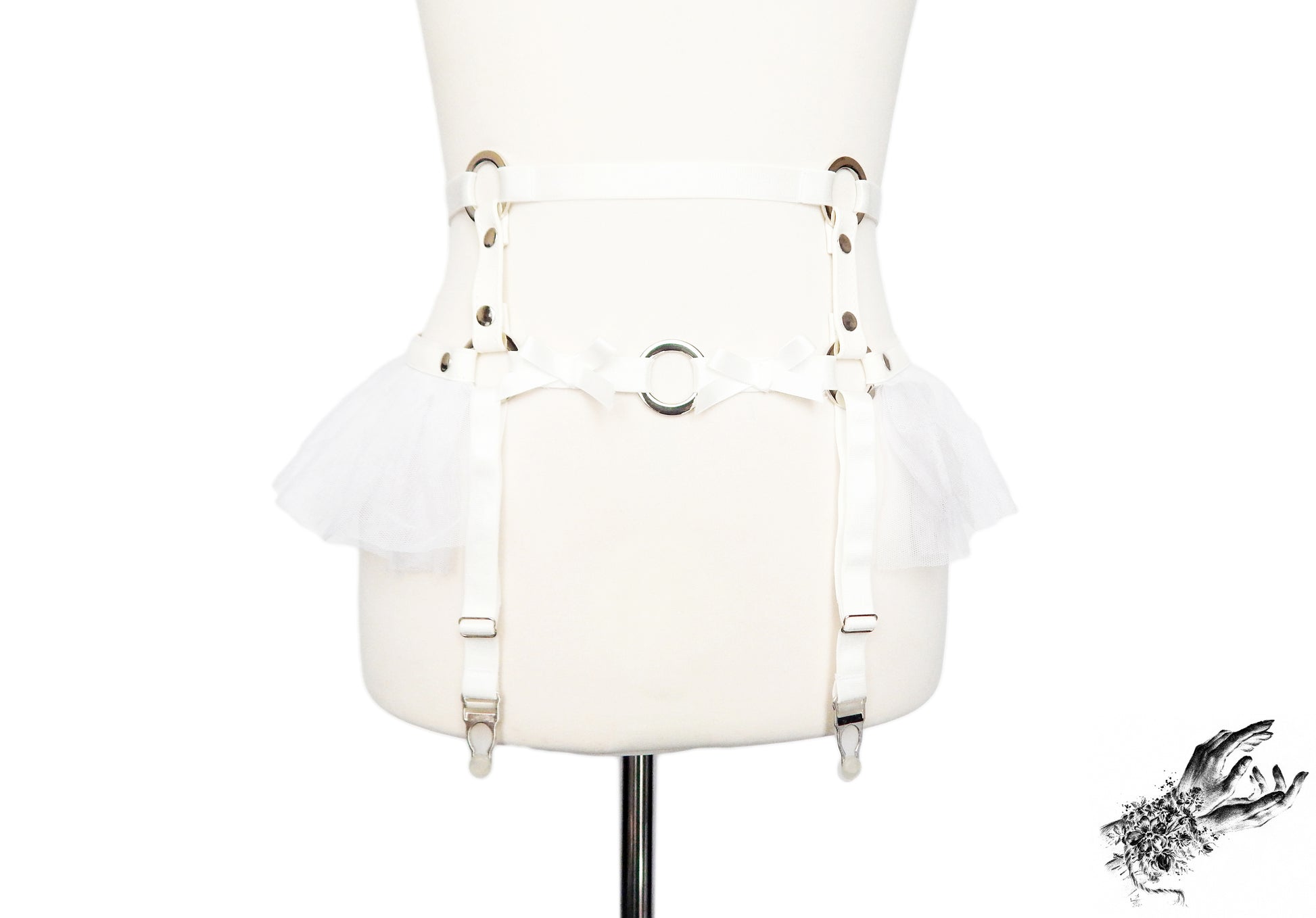 White faux leather garter belt featuring silver O ring hardware, white ruffled tulle and bow appliques at the waist with adjustable suspender straps, displayed on a mannequin against a white background.