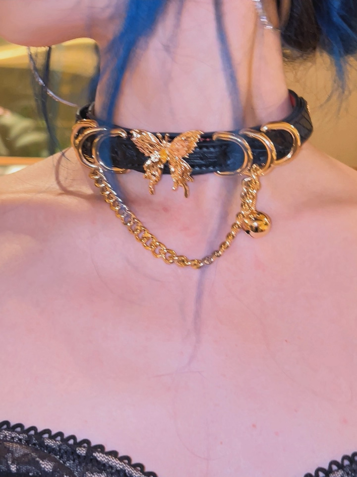 Worn photo of a black and red leather choker featuring a gold tone butterfly charm, D rings, chain and bell charm. Styled with black and blue hair and a black lace top for a glamorous gothic aesthetic.