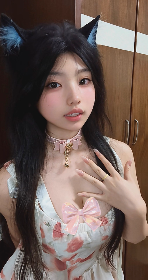 Worn photo of a pink leather choker featuring a central bow shaped design, gold tone rose and heart studs with detachable pink and gold crystal heart and bell charm. Styled withblack animal ears and a white and pink babydoll dress for a kawaii fashion aesthetic.