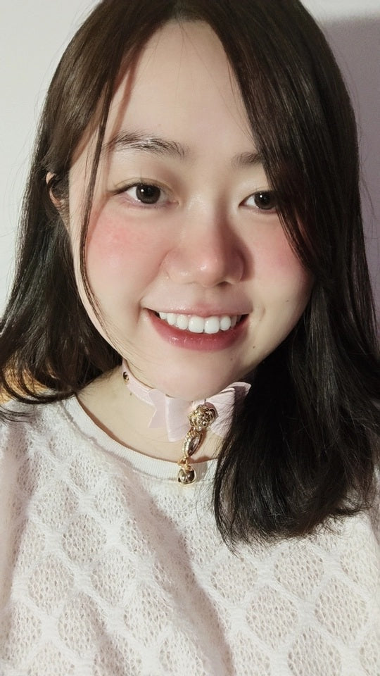 Worn photo of a pink leather choker featuring a central bow shaped design, gold tone rose and heart studs with detachable pink and gold crystal heart and bell charm. Styled with soft blush pink make-up and a pastel pink t-shirt for a Larme Kei fashion aesthetic.