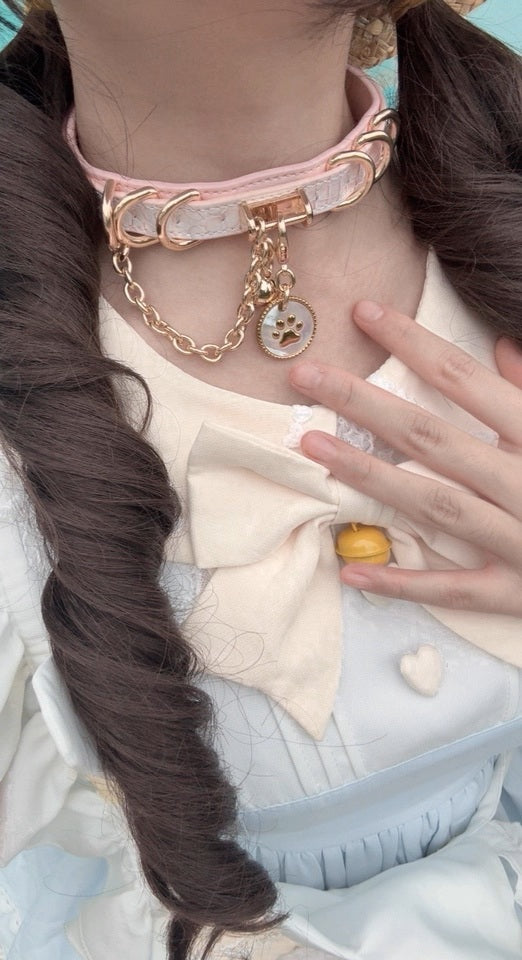 Worn photo of a pink leather choker featuring a white and gold paw print pet tag, gold tone chain, D rings and bell charm. Styled with brown curly pigtails and a baby blue and white maid costume for a kawaii idol aesthetic.