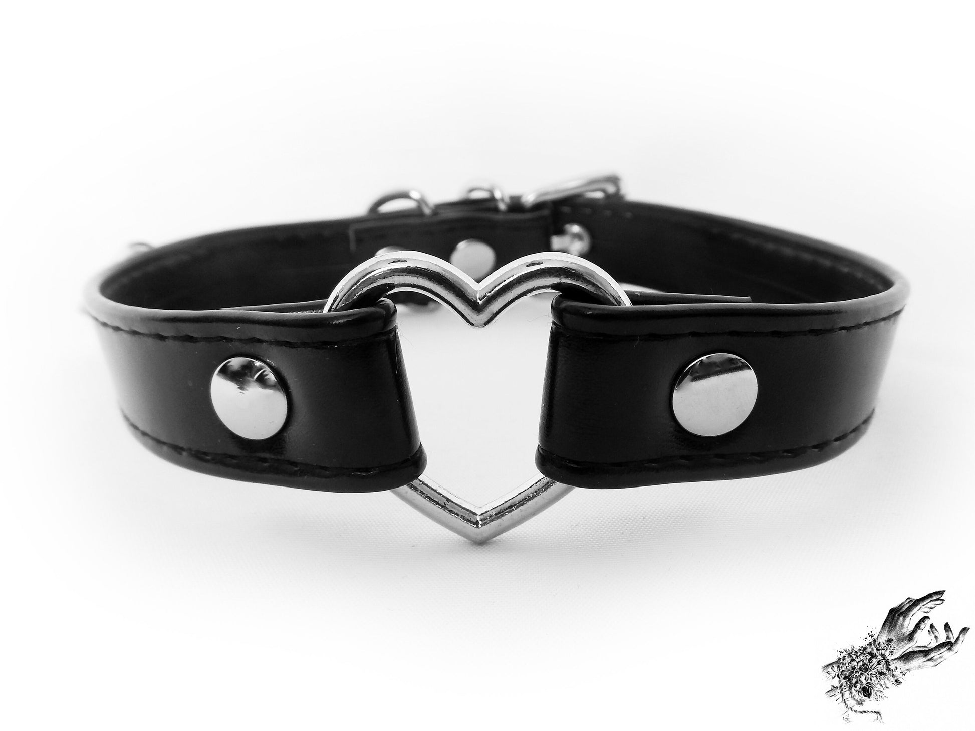 Close-up of a black faux leather choker featuring a central silver tone heart ring, stainless steel plated hardware and adjustable buckle fastening at the back. Displayed on a white background.