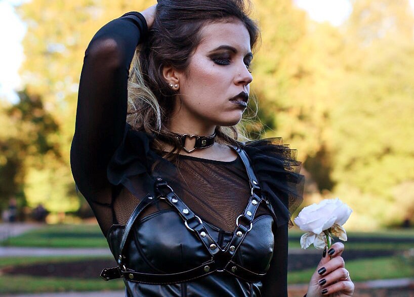 Modelled photo of a black faux leather choker featuring a central silver tone heart ring and stainless steel plated hardware. Styled with a black faux leather harness, mesh shirt and black lipstick for a gothic fashion style.