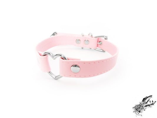Pink faux leather choker featuring a central silver tone heart ring, stainless steel plated hardware and adjustable buckle fastening at the back. Displayed against a white background.