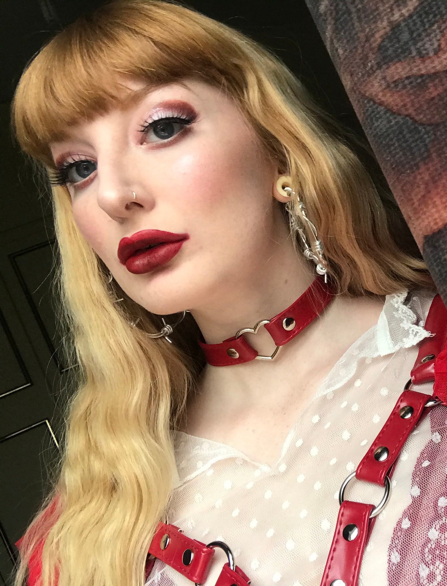 Modelled photo of a red faux leather choker featuring a central silver tone heart ring and stainless steel plated hardware, with a buckle fastening and multiple sizing holes at the back. Styled with a red faux leather harness, sheer white polka dot blouse and red lipstick for a an alternative vintage look.