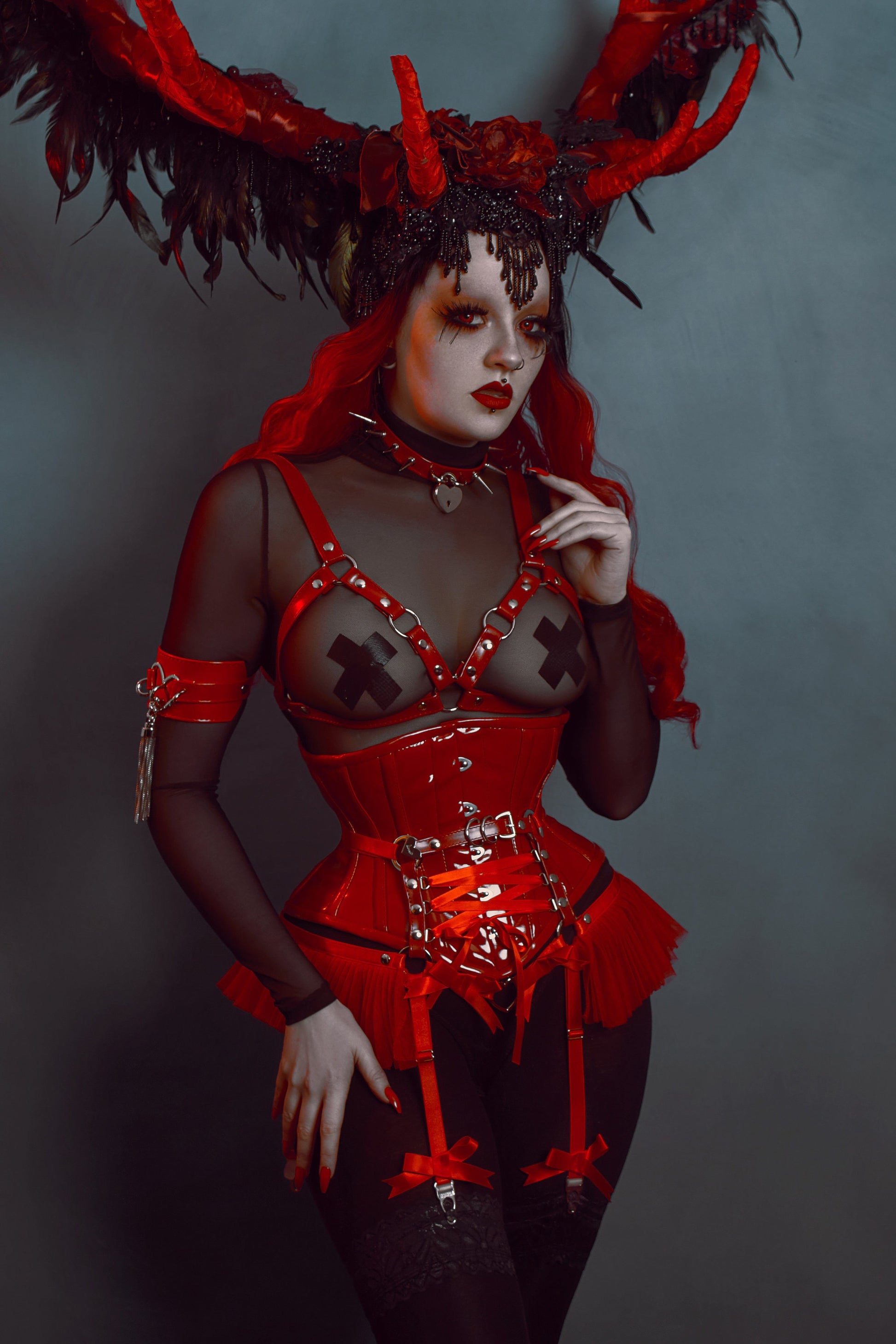 Modelled photo of a red vegan leather harness set featuring silver O ring hardware, red ruffled tulle and corset lacing at the waist, styled with a sheer black mesh top and red PVC corset for a gothic look.
