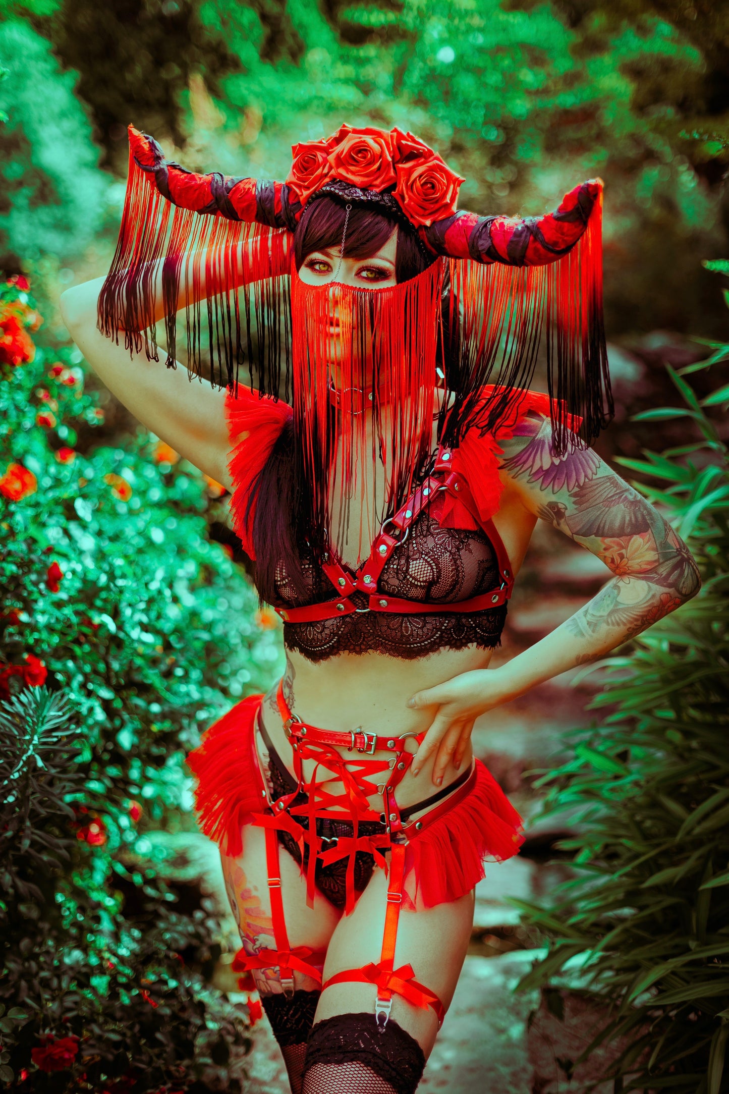 Modelled photo of a red vegan leather harness set featuring ruffled tulle accents at the shoulders and waist with ribbon corset lacing, styled with black lace lingerie and an elaborate black and red fringed headpiece for a gothic fantasy look.