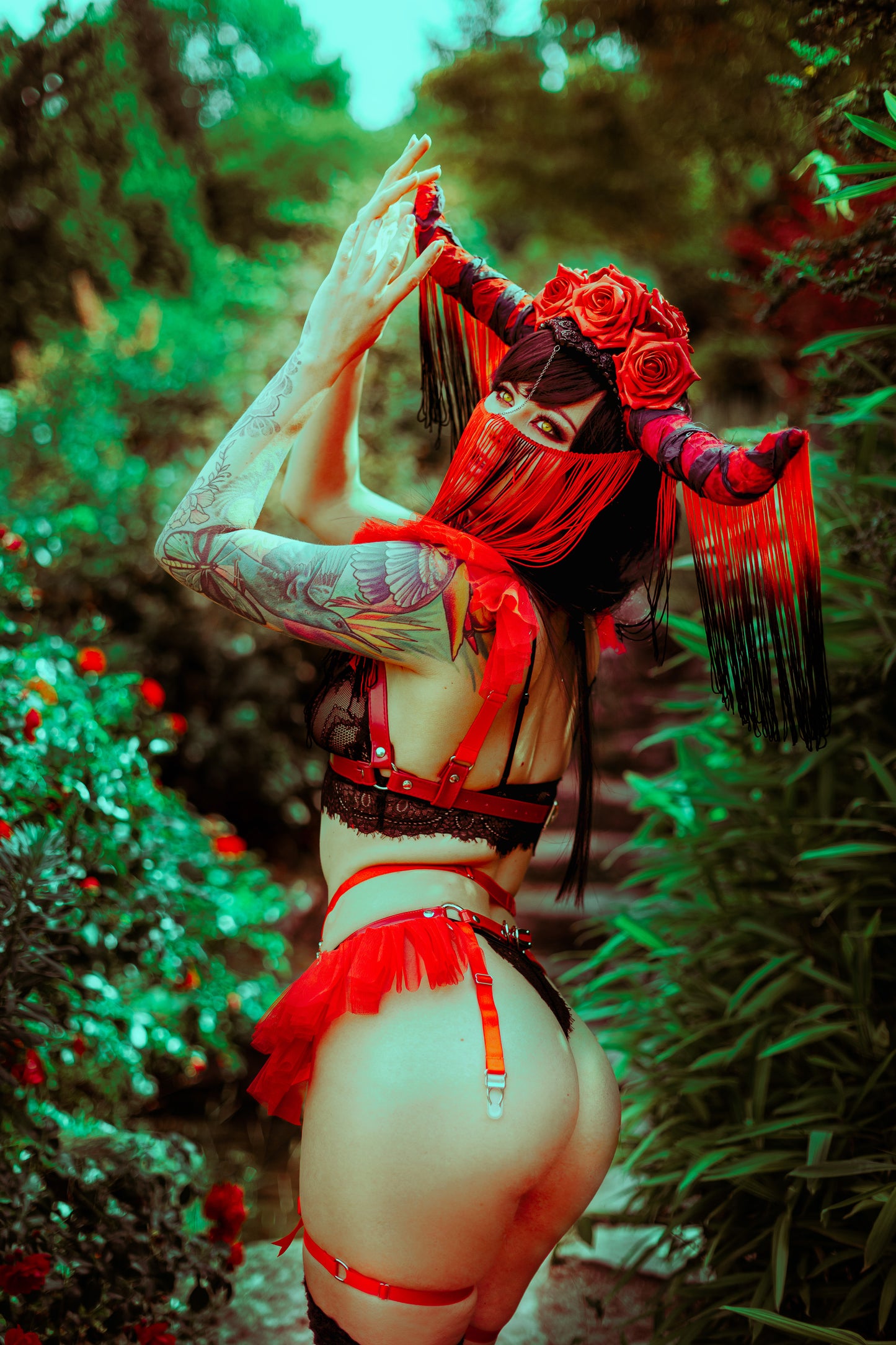 Rear view modelled photo of a red vegan leather harness set showing the adjustable elastic and buckled faux leather body straps, styled with black lace lingerie and an elaborate black and red fringed headpiece for a gothic fantasy look.