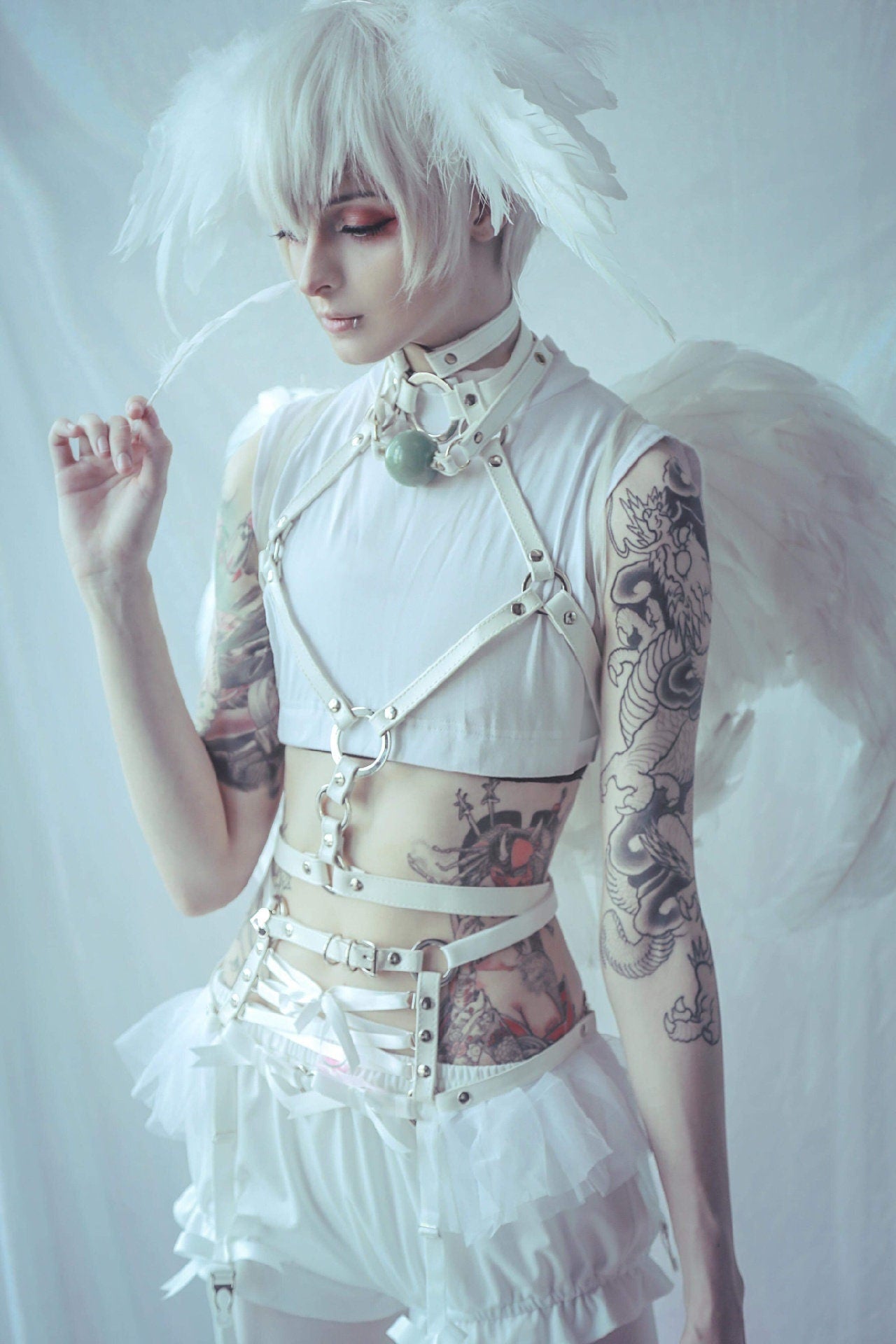 Modelled photo of a white faux leather harness set featuring silver O ring hardware. white ruffled tulle and corset lacing at the waist, styled with white crop top, ruffled bloomers and feathered wings for a kawaii angel aesthetic.