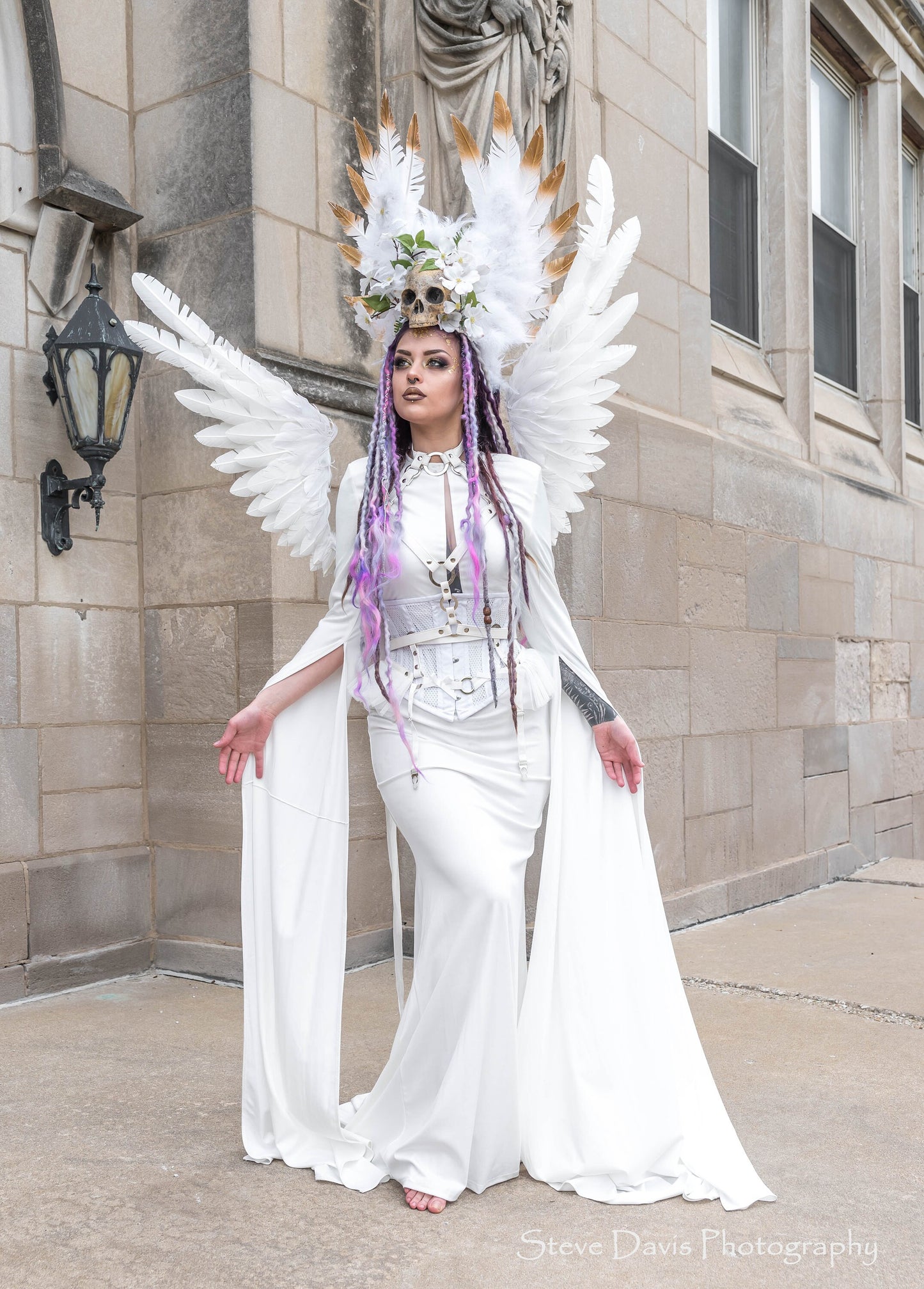 Modelled photo of a white vegan leather harness set featuring silver O ring hardware and white ruffled tulle accents at the waist, styled with an elaborate skull headpiece and feathered wings for a dark angel look.